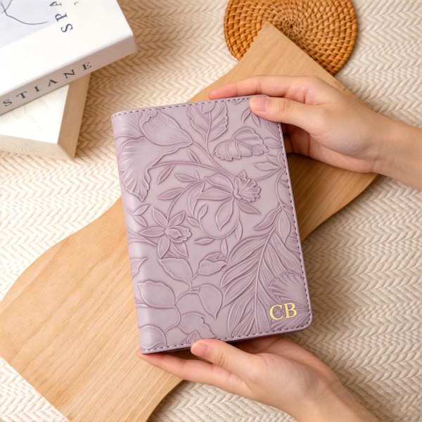 May include: A rectangular, light purple passport holder featuring an embossed floral pattern across the front surface. The bottom right corner displays the gold-colored, capitalized initials CB. The item is held in a vertical orientation.