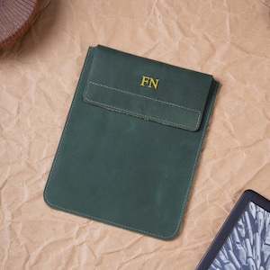 May include: A dark green leather sleeve with a flap closure and the letters FN embossed in gold on the front. The sleeve has a rectangular shape with rounded bottom corners and visible stitching along the edges, providing a secure storage pouch.