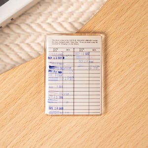 May include: A vintage library card in a clear plastic sleeve, with handwritten entries and stamped dates. The card is on a light wood surface, with a glimpse of a book and a woven basket in the background.