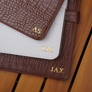 May include: Three rectangular folders with a textured crocodile skin pattern. Two folders are brown and one is white. Each folder features gold embossed lettering: the top brown folder has AS, the white folder has KM, and the bottom brown folder has JAX.