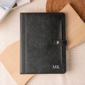 May include: A black leather portfolio with a snap closure and stitched edges. The initials "MK" are visible in the lower right corner. The portfolio is resting on a light-colored surface.