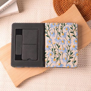 May include: Open, dark gray leather journal with a floral embroidered cover featuring blue flowers and green stems. The left side has pockets for cards and a pen. The journal is open on a wooden cutting board.