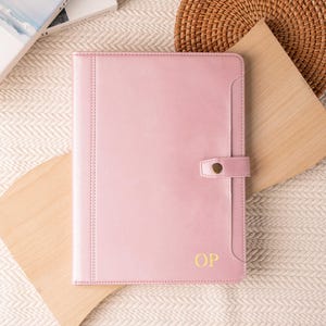 Könnte beinhalten: Ein hellrosa Lederjournal oder Portfolio mit Schnappverschluss. Der Einband trägt die goldenen Initialen "OP". Das Journal liegt auf einer hellen Oberfläche mit Holzakzenten und einem geflochtenen Korb.