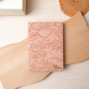 Puede incluir: Un portapasaporte de color rosa claro con un patrón floral en relieve en toda su superficie. En la esquina inferior derecha de la cubierta aparece el nombre Yan en una escritura cursiva oscura y elegante, añadiendo un toque personalizado.