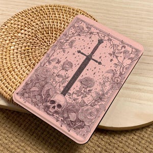 Peut inclure: Un &eacute;tui rectangulaire rose avec une illustration d&eacute;taill&eacute;e d'une &eacute;p&eacute;e, d'un cr&acirc;ne, de roses et de vignes. L'&oelig;uvre d'art est dans des tons de gris et de marron, sur un fond rose. L'&eacute;tui est pos&eacute; sur une surface tiss&eacute;e.