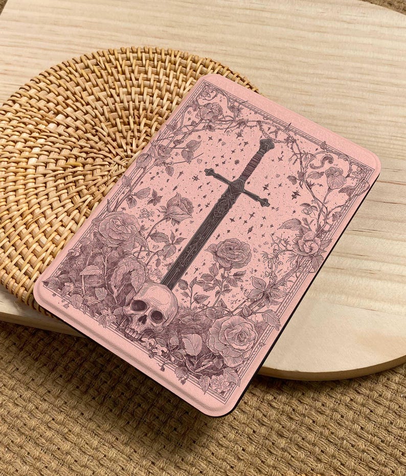 Peut inclure: Un &eacute;tui rectangulaire rose avec une illustration d&eacute;taill&eacute;e d'une &eacute;p&eacute;e, d'un cr&acirc;ne, de roses et de vignes. L'&oelig;uvre d'art est dans des tons de gris et de marron, sur un fond rose. L'&eacute;tui est pos&eacute; sur une surface tiss&eacute;e.