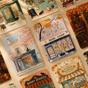 May include: A collection of rectangular e-reader cases featuring various illustrated storefronts, including a book shop with pink trees, a pumpkin bookshop, and a spooky dessert shop, all displaying detailed architectural designs and seasonal themes.