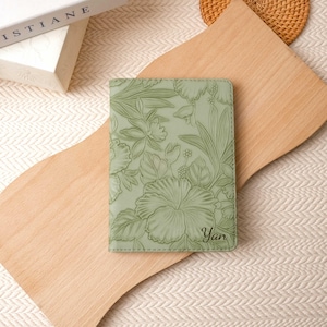 May include: A sage green passport holder with a floral embossed design featuring hibiscus flowers and foliage. The name "Yan" is printed in the lower right corner. The passport holder is on a wooden cutting board.