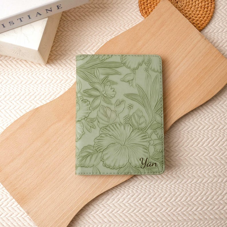 May include: A sage green passport holder with a floral embossed design featuring hibiscus flowers and foliage. The name "Yan" is printed in the lower right corner. The passport holder is on a wooden cutting board.