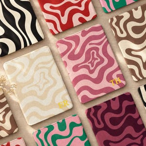 May include: A collection of rectangular tablet or e-reader sleeves featuring wavy, swirling patterns in various color combinations including black and white, red and cream, pink and magenta, and green and pink, with some featuring gold initials.