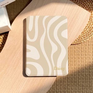 Può includere: Un taccuino beige e crema con un motivo a onde. La parola "Daisy" è stampata in oro nell'angolo in basso a destra. Il taccuino è rettangolare e si trova su una superficie di legno.