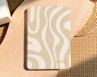 May include: A rectangular e-reader case featuring a wavy, abstract pattern in shades of beige and cream. The surface has a smooth finish with the name Daisy printed in small, gold-colored serif lettering near the bottom right corner.