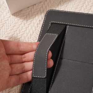 May include: A black tablet case with a hand strap and white stitching. The case has a built-in elastic band and a pocket. The word "MINIMAL" is visible on a white box in the background.
