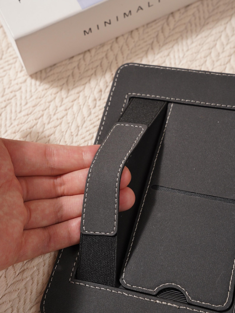 May include: A black tablet case with a hand strap and white stitching. The case has a built-in elastic band and a pocket. The word "MINIMAL" is visible on a white box in the background.