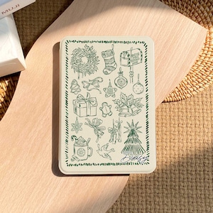 May include: A rectangular, off-white Christmas-themed card with a green border and illustrations. The card features drawings of a wreath, stocking, mailbox, ornaments, gifts, gingerbread man, bells, and a Christmas tree. The name "Daisy" is written in cursive.