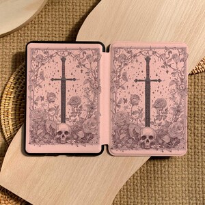 Peut inclure: Un &eacute;tui rose pour liseuse &eacute;lectronique avec un design gothique. L'&eacute;tui pr&eacute;sente une &eacute;p&eacute;e noire, des roses, des cr&acirc;nes et des vignes. Le design est sur fond rose. L'&eacute;tui est ouvert, r&eacute;v&eacute;lant l'int&eacute;rieur.