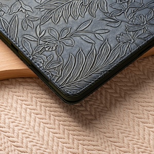 May include: A close-up of a dark blue leather laptop case with a floral embossed design. The case has a stitched edge and is lying on a light beige surface.