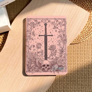 Peut inclure: Un &eacute;tui rectangulaire rose avec un motif d'&eacute;p&eacute;e noire entour&eacute;e de roses, de vignes et d'un cr&acirc;ne. Le mot "Daisy" est &eacute;crit en cursive. L'&eacute;tui est pos&eacute; sur une surface en bois clair.