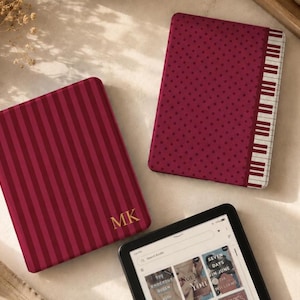Może przedstawiać: Dwa etui na czytnik e-booków w kolorze ciemnoczerwonym, jedno w pionowe paski ze złotymi literami monogramu MK, a drugie w kropki z obwódką w kształcie klawiszy pianina, obok czytnika wyświetlającego cyfrową bibliotekę okładek książek.