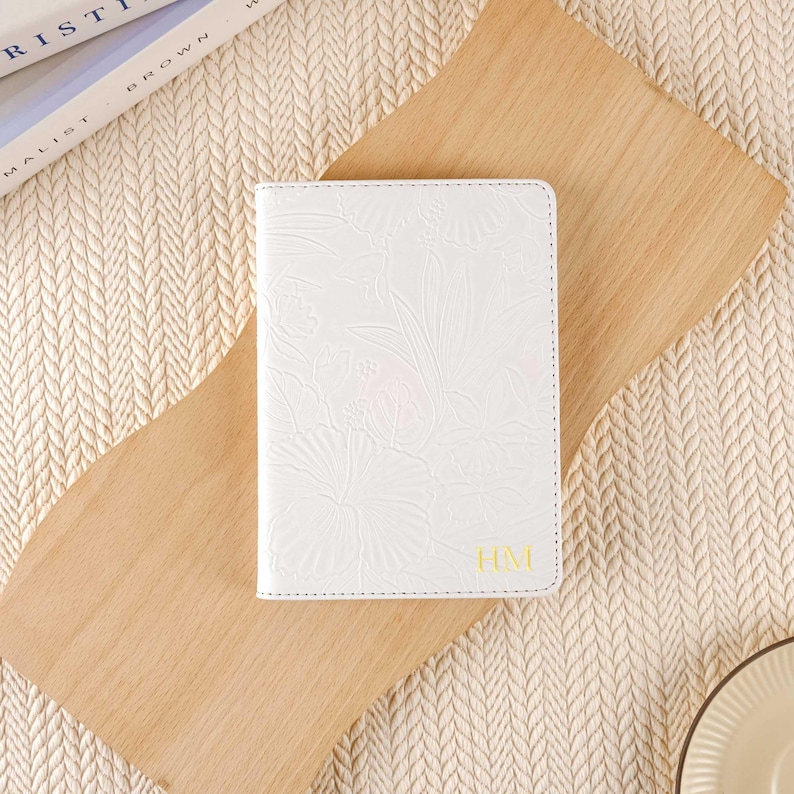 May include: White leather passport holder with an embossed floral design and the initials "HM" in gold lettering.