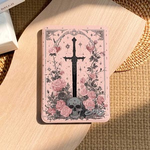 以下が含まれることがあります： バラ、心臓、頭蓋骨に囲まれた黒い剣のデザインが特徴のピンク色の長方形ケース。「Daisy」の文字が筆記体で書かれています。ケースには装飾的なボーダーがあり、明るい木製の表面に置かれています。