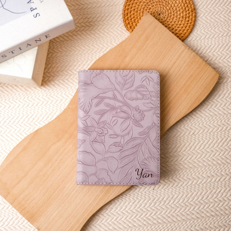 May include: A lavender passport holder with an embossed floral design. The cover features a detailed pattern of flowers and leaves. The name "Yan" is inscribed in a script font on the lower right corner.