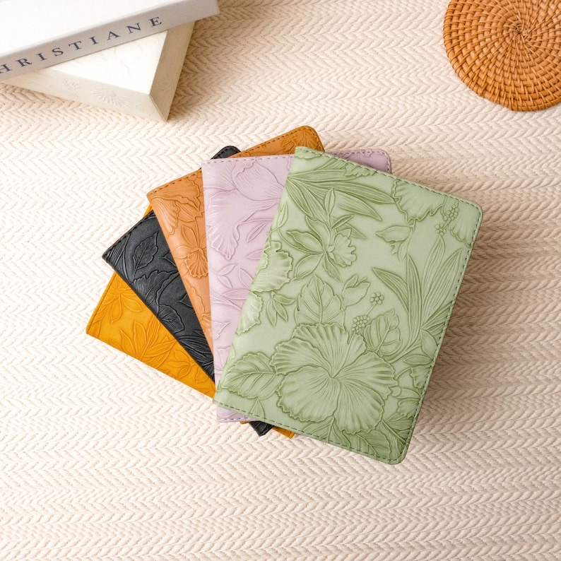 May include: A collection of five leather journals in various colors: yellow, black, orange, lavender, and green. Each journal features a floral embossed design. The journals are arranged in a fan shape, with the green journal in the foreground.