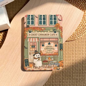 Może przedstawiać: Prostokątne etui na tablet z kolorową ilustracją witryny kawiarni o nazwie Ghost Cinnamon Cafe. Scena przedstawia ducha w kapeluszu czarownicy, dynie, jesienne liście, girlandy świetlne oraz meble kawiarniane w jesiennym klimacie.