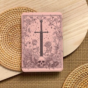 Peut inclure: Une bo&icirc;te rectangulaire rose avec une illustration d&eacute;taill&eacute;e d'une &eacute;p&eacute;e, de roses, d'un cr&acirc;ne et d'&eacute;toiles. L'&oelig;uvre d'art est de style vintage, avec une &eacute;p&eacute;e sombre au centre et des &eacute;l&eacute;ments floraux. La bo&icirc;te est pos&eacute;e sur une surface en bois.