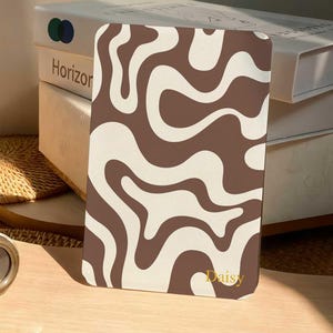 May include: A rectangular flat object featuring a brown and cream wavy, abstract pattern across its surface. The name Daisy is printed in small, gold-colored lettering near the bottom right corner of the item.