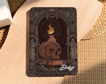 May include: A dark brown and gold illustrated card with a skull on top of a book. The skull is lit by a flame and the card has an ornate border. The text "Daisy" is written in cursive at the bottom of the card.