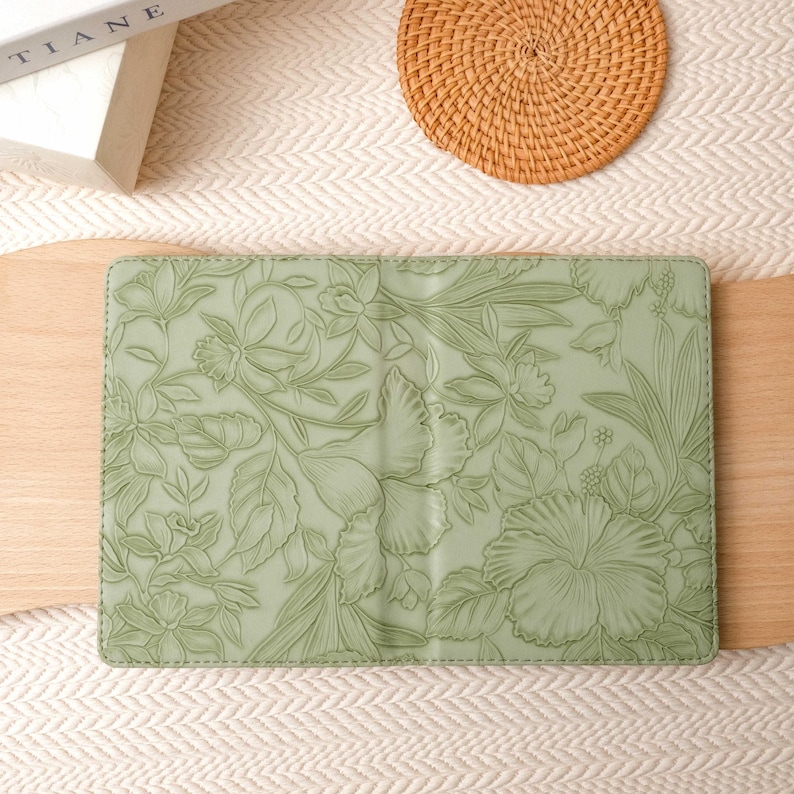 May include: A sage green journal cover with an embossed floral design featuring hibiscus and other tropical flowers. The cover is lying on a wooden surface next to a woven coaster and a book.