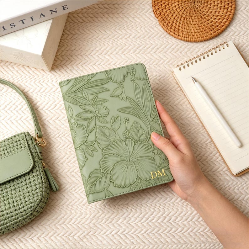 May include: A sage green journal with embossed floral designs and gold initials. The journal is held by a person's hand. A matching green handbag, a notepad with a pen, and a woven coaster are also in the image.