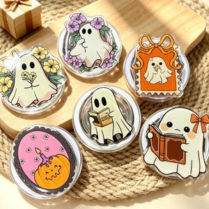 May include: Six circular acrylic phone grips featuring ghost illustrations. One ghost holds yellow flowers, one is surrounded by purple flowers, one is in an orange stamp frame, one reads a book, one reads a brown book with a bow, and one is a pumpkin.