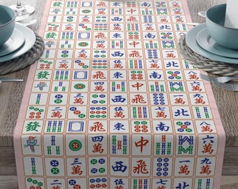 May include: A rectangular table runner featuring a Mahjong tile pattern in various colours on a light pink background. The runner is laid on a wooden table set for a meal, with plates, bowls, and wine glasses.