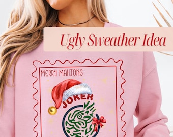 May include: A light pink sweatshirt with a red and white graphic that reads "Merry Mahjong" and "Joker" with a Santa hat. The graphic is framed in a red postage stamp design. The text "Ugly Sweater Idea" is above the graphic.