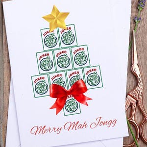 May include: A Christmas card with a tree made of Joker playing cards, topped with a gold star and a red bow. The card reads "Merry Mah Jongg" in red script. The card is in a wooden tray with scissors and lavender.