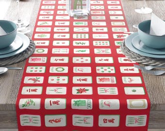 May include: A red table runner with a repeating pattern of white rectangular tiles featuring Chinese characters and festive illustrations. The runner is set on a wooden table, with place settings and a vase of pink lilies.