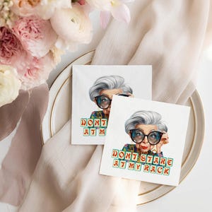 Op de afbeelding: Twee vierkante witte servetten met een kleurrijke illustratie van een vrouw met een grote bril en de tekst "DON'T STARE AT MY RACK". De servetten liggen op een witte plaat met een gouden rand, met bloemen en een beige doek op de achtergrond.