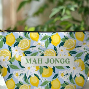 May include: A zippered pouch with a light blue background, featuring a pattern of yellow lemons, white flowers, and green leaves. The text "MAH JONGG" is displayed on a rectangular label. The pouch has a gold zipper.