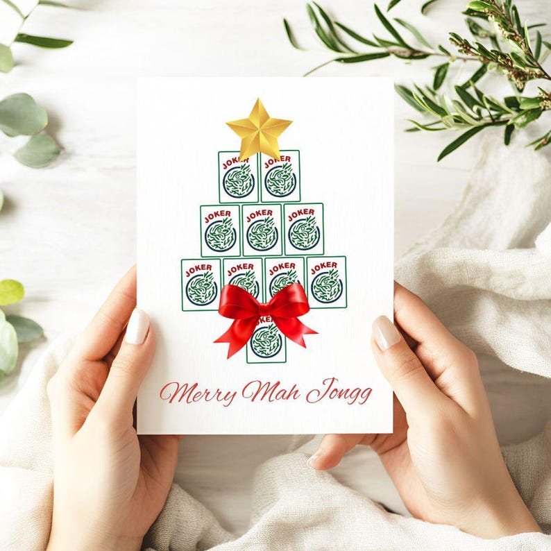 May include: A white greeting card featuring a Christmas tree design made of Mahjong tiles. The tree is topped with a gold star and adorned with a red bow. The text "Merry Mah Jongg" is written in red script below the tree.