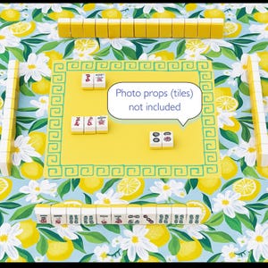 Puede incluir: Un tapete de juego de Mahjong amarillo con un borde verde, rodeado de fichas de Mahjong. El tapete est&aacute; sobre un fondo azul con un patr&oacute;n de lim&oacute;n y flores. El texto "Photo props (tiles) not included" est&aacute; en un bocadillo blanco.