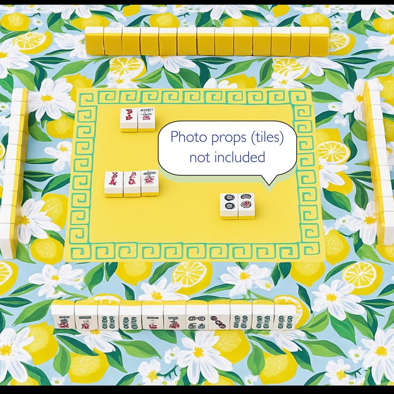 Puede incluir: Un tapete de juego de Mahjong amarillo con un borde verde, rodeado de fichas de Mahjong. El tapete est&aacute; sobre un fondo azul con un patr&oacute;n de lim&oacute;n y flores. El texto "Photo props (tiles) not included" est&aacute; en un bocadillo blanco.