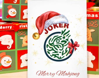 Könnte beinhalten: Eine weiße Grußkarte mit einem roten Weihnachtsmannhut mit dem Wort "JOKER" über einem grünen und blauen kreisförmigen Design. Die Karte trägt die Aufschrift "Merry Mahjong" in roter Schrift. Weihnachtsgeschenkboxen sind im Hintergrund.