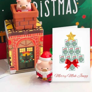 May include: A Christmas-themed scene featuring a pig figurine in a Santa suit, a pig in a chimney, and a greeting card. The card displays a Christmas tree made of Joker playing cards, a red bow, and the text "Merry Mah Jongg".