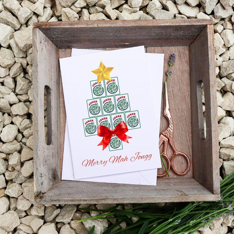 May include: A Christmas card with a tree made of Joker playing cards, topped with a gold star and a red bow. The card reads "Merry Mah Jongg" in red script. The card is in a wooden tray with scissors and lavender.