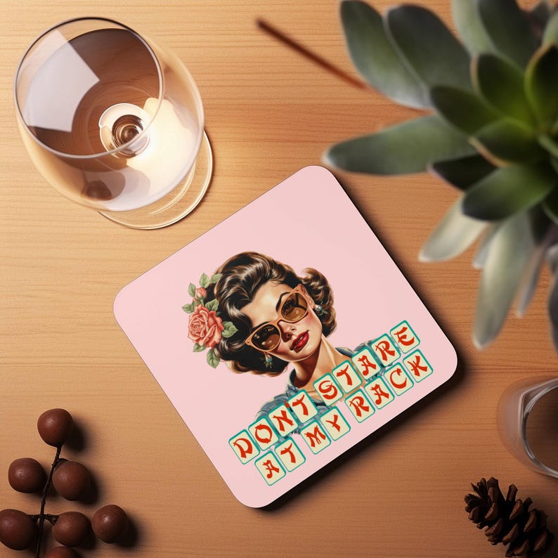 May include: A square pink coaster featuring a vintage illustration of a woman with sunglasses and a flower in her hair. The coaster has the text "DON'T STARE AT MY RACK" in a retro font. A glass of wine is visible in the background.