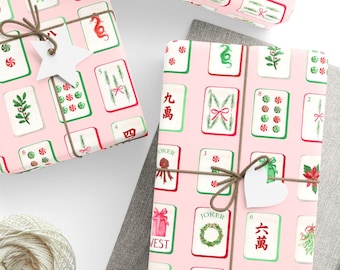 May include: Gift-wrapped presents with pink wrapping paper featuring a Mahjong tile design in red, green, and white. The gifts are tied with twine and adorned with white star and heart-shaped tags. A spool of twine and gray fabric are also visible.
