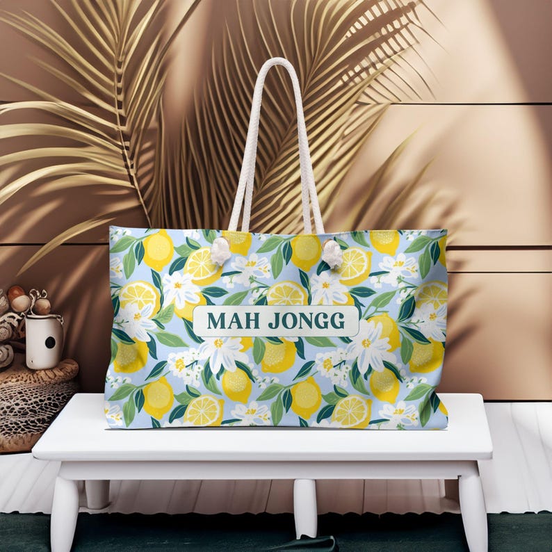 Pu&ograve; includere: Una grande borsa tote con sfondo azzurro, con un motivo di limoni gialli, fiori bianchi e foglie verdi. La borsa ha manici in corda bianca e il testo "MAH JONGG".