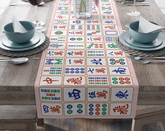 May include: A pink table runner with a repeating pattern of Mahjong tiles in various colours. The tiles display Chinese characters and symbols. The runner is set on a wooden table, with place settings and a vase of pink flowers.